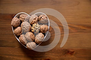 Whole walnuts and one cracked broken heart in a white heart-shaped bowl on a wooden background. Top view. Place for text