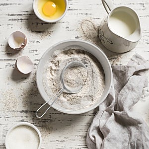 Whole-wheat flour in white bowl, milk, eggs on a light wooden background.