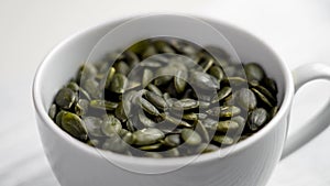 Wholesome green pumpkin seeds falling into a white ceramic cup.