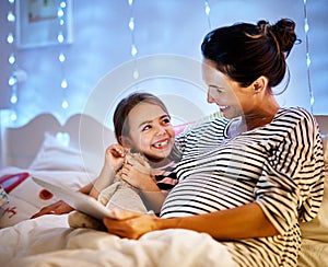 Whos picking the story tonight. an attractive young pregnant woman reading her daughter a bedtime story on a tablet.