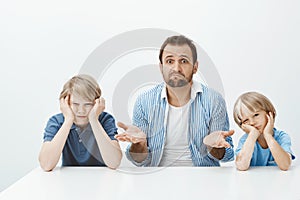 Why sons are mad. Portrait of clueless nervous father-in-law sitting with two cute boys at table, shrugging and being