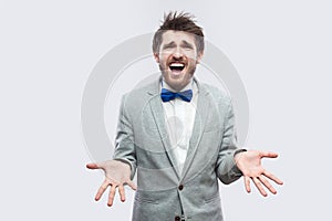 Why? what? Portrait of confused handsome bearded man in casual grey suit, blue bow tie standing and looking at camera with raised