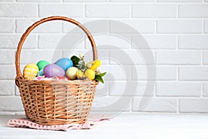 Wicker basket with bright painted Easter eggs and spring flowers on white wooden table near brick wall. Space for text