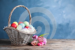 Wicker basket with bright painted Easter eggs and spring flowers on wooden table against blue background. Space for text
