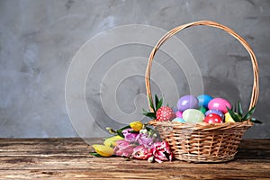 Wicker basket with bright painted Easter eggs and spring flowers on wooden table against grey background. Space for text