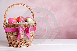 Wicker basket with bright painted Easter eggs on white wooden table against pink background. Space for text