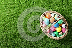 Wicker basket with Easter eggs on green grass, top view. Space for text