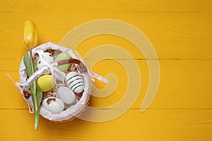 Wicker basket with festively decorated Easter eggs and beautiful tulip on yellow wooden background, top view. Space for text