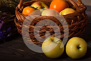 A wicker basket filled with fruits, persimmons and apples, in front of the basket lies two apples.