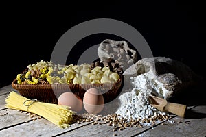 Wicker Basket Filled with Pasta of Different Colors and Shapes, Flour in Jute Bag and Two Eggs on Wooden Table On Black