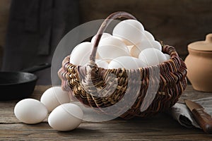 Wicker basket full of white chicken eggs on table. Frying pan and clay pot on background.