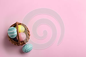 Wicker basket with painted Easter eggs on color background, top view.