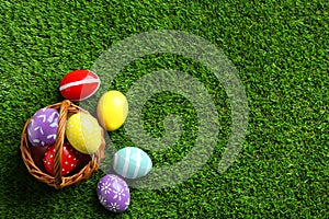 Wicker basket with painted Easter eggs on green grass, top view.