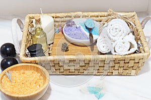 Wicker basket with a set for spa treatments, colorful salt, aromatic oil, stones, candle and soft towels, next to a white table Bi