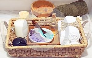 Wicker basket with a set for spa treatments, multi-colored salt, aromatic oil, stones, candle and soft towels, next to a white tab