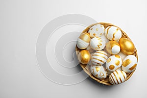 Wicker basket of traditional Easter eggs decorated with golden paint on white background, top view.
