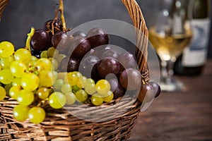 Wicker basket with various grapes: red, white and black berries on the dark wooden table with bottle and glass of white wine in th