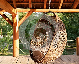 A wicker chair with cushions is suspended from the strong wooden beams of the veranda against the background of green trees.