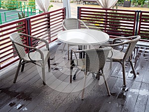 Wicker chairs stand around a white round table on a wooden floor wet after rain in an outdoor cafe