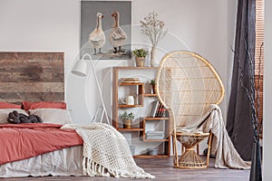 Wicker peacock chair with blanket in fashionable rustic bedroom with bookshelf and king size bed with bedhead