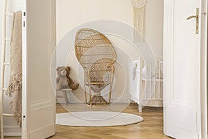 Wicker peacock chair standing in white baby room interior with crib, teddy bear and round rug on herringbone parquet