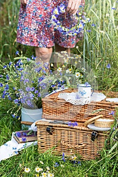 Wicker picnic suitcases with cup on it in wheat field and woman holding wreath of cornflowers and chamomile