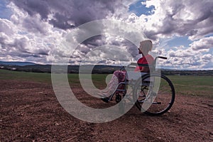 Wide angle disabled girl sitting in a wheelchair in the middle of a green field in the countryside with a sky clouds