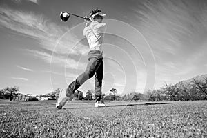 Wide angle grayscale shot of a Caucasian male golfer teeing off from a golf tee on a sunny day