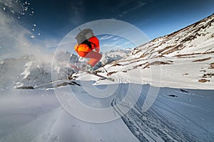 Wide angle male skier in orange suit makes a jump from a snowy ledge in the mountains. Snow powder trailing behind the