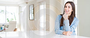 Wide angle picture of beautiful young woman sitting on white table at home looking confident at the camera with smile with crossed