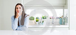 Wide angle picture of beautiful young woman sitting on white table at home looking confident at the camera with smile with crossed