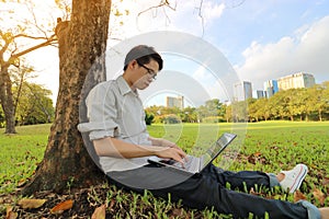 Wide angle shot of happy young man typing on a laptop computer for his work in a beautiful city park.