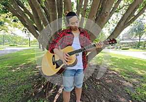 Wide angle shot of portrait of handsome young man playing acoustic guitar in the park outdoors with a large tree background.