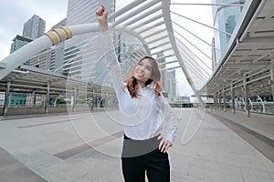 Wide angle shot of successful young Asian business woman raising her hand and smiling at urban building background