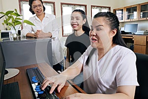 Wide angle view of happy Asian women co-workers in office workplace including person with blindness disability using computer with photo