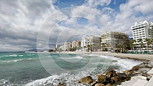 Wide-angle view of a seaside cityscape under dynamic skies, flanked by lush palm trees and framed by the Mediterranean