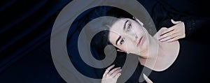 Wide banner panorama portrait of black haired young lying woman with beguiling hypnotic look on blue black fabric background,