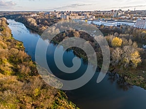 A wide and calm river flows through a large city, past a park and a forest. Autumn landscape. View of the river from a drone.