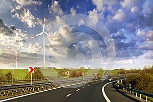 Wide highway with moving car stretching to horizon by green hills with high wind turbines on blue cloudy sky background. Modern te