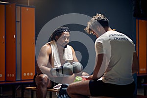 Wide horizontal shot of strong muscular boxing player after workout in sweats resting on bench with coach