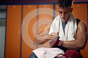 Wide horizontal shot of young sweaty man taking red protective tape of his hands after boxing workout