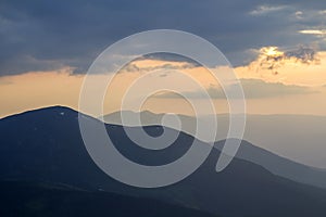 Wide panorama, fantastic view of covered with morning mist green Carpathian mountains at dawn under dark clouds and light pink sky