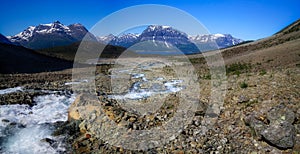 A wide panorama of the mountain landscape with snowy peaks, a bubbling mountain river flowing down the valley among stones and