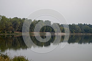 Wide river flowing across green forest. Fall. Evening. Reflections of trees in the calm water. Sundown