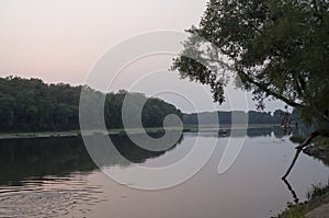 Wide river flowing across green forest. Fall. Evening. Reflections of trees in the calm water. Sundown