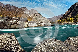 Wide river flows along stony banks among rocky mountains against clear blue sky. Turquoise water of stormy river and huge stones.