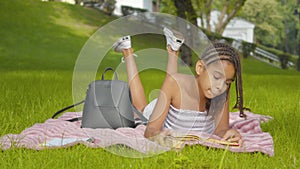 Wide shot of absorbed concentrated girl lying on green meadow in park and reading book. Portrait of charming African