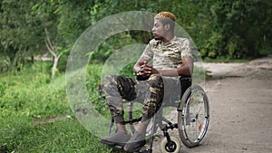 Wide shot African American young military man in wheelchair looking away admiring beauty of summer nature in parkland