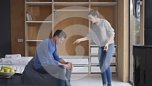 Wide shot angry aggressive woman shouting gesturing as man sitting on bed at home. Side view portrait furious Caucasian