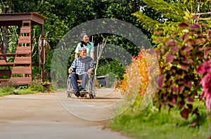 Wide shot of Asian nurse cart of shove senior man sit on wheelchair to walk along the walkway in the garden and they look